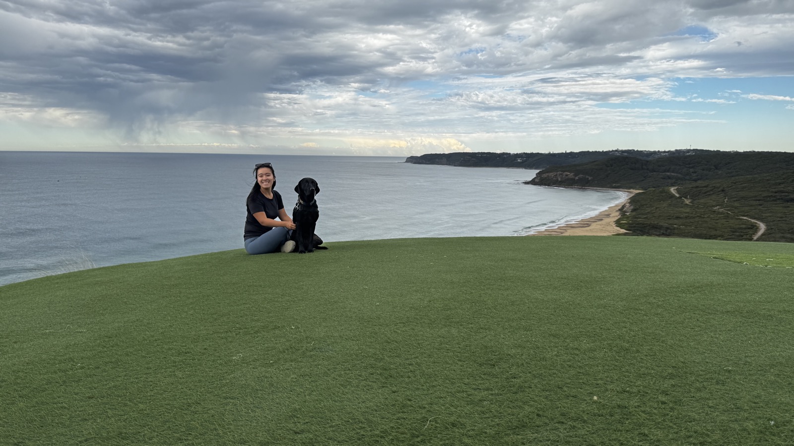 Diona and Murphy the black Labrador sitting together on a clifftop lawn above the ocean, no lead in anyone's hand