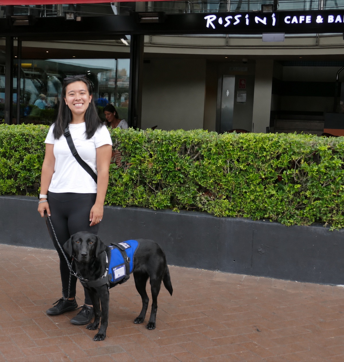 Diona standing outside a cafe with Murphy, her black Labrador assistance dog, calm in her blue working vest